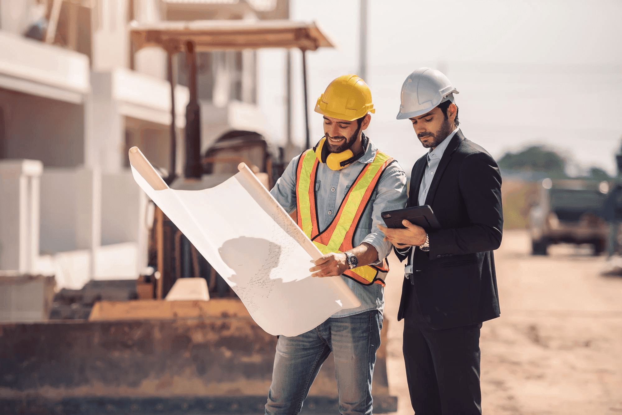 Two construction professionals review building plans at a sunny outdoor construction site.