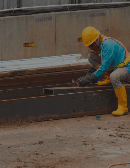 Construction worker in yellow hard hat kneeling, using a tool on a metal beam.