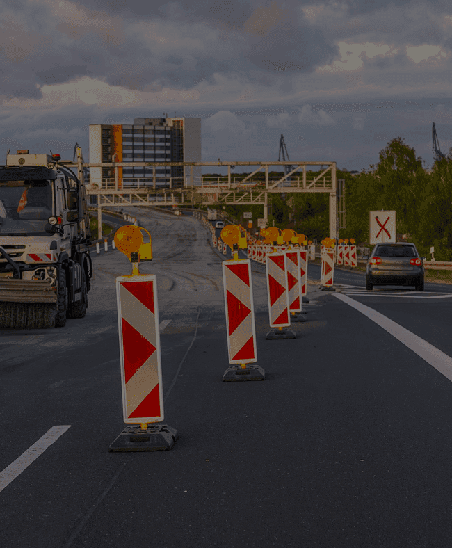 Road under construction with orange and white traffic barriers, a truck, and cars.