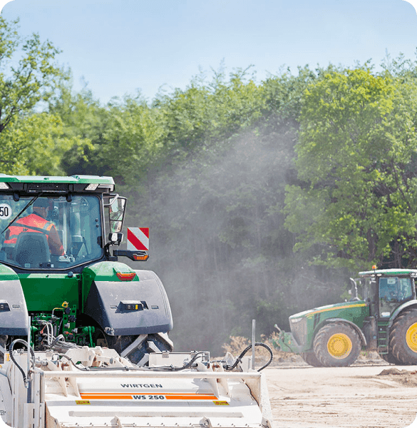 Two green John Deere tractors operate on a dusty construction or agricultural site.