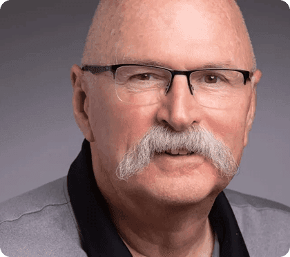 Bald man with a prominent white mustache, black-rimmed glasses, and grey collared shirt.
